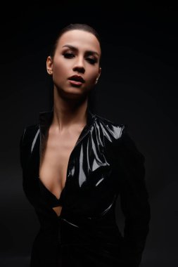 Bir kadın, kendine güvenen siyah bir elbise içinde, güzelliğini ve duruşunu sanatsal bir ortamda ön plana çıkarıyor..