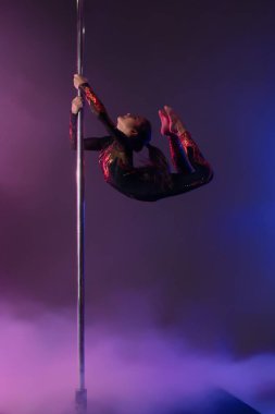 Bir kız striptiz dans yeteneğini sergiliyor, zarif bir şekilde direğin yanında asılı duruyor yumuşak ışıklarla renkli bir ortamda..
