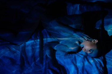 Bir kadın, yansıyan ışıkla üstsüz poz verir. Cildine canlı gölgeler bırakır. Sanatçılık ve samimiyet hissi uyandırır..