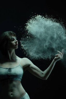 Bir dansçı zarifçe etrafında dönen toz parçacıklarıyla poz verir, karanlık bir ortamda sanatçılık ve hareket sergiler..