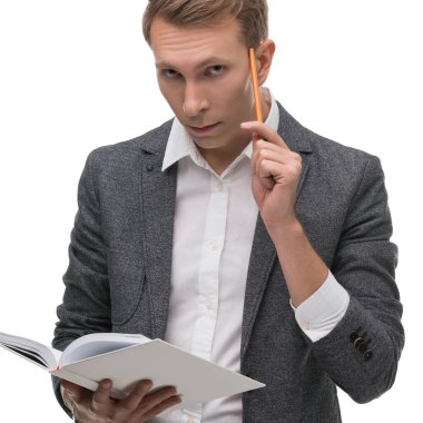 Gri takım elbiseli bir adam elinde kalemle düşünürken bir kitap tutar, düz bir arkaplanda derin düşüncelere dalmış görünür..