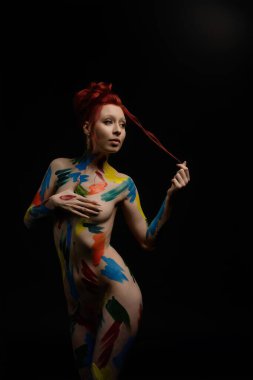 Bir kadın zarif bir şekilde ayakta duruyor, karanlık bir arka plana karşı canlı renklerle süslenmiş vücut sanatını sergiliyor. Sanat eserleri onun güzelliğini ve formunu vurguluyor..