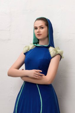Genç bir manken şık bir şekilde kollarını kavuşturmuş, kapüşonlu ve zarif omuz detaylarıyla basit bir stüdyoda mavi bir elbise giyiyor..