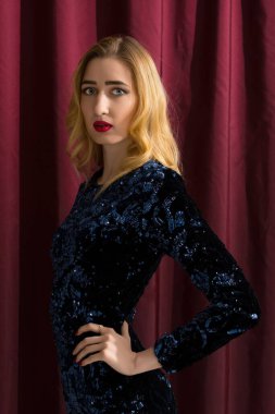 Bir model parlak, koyu renk bir elbisenin içinde duruyor, şık görünüşünü ve çarpıcı özelliklerini koyu bir bordo perdeye karşı sergiliyor..