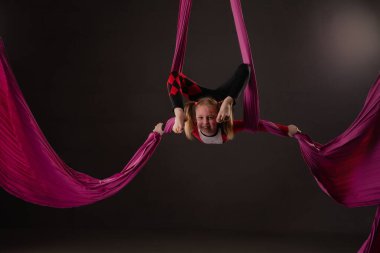 Genç bir kız, stüdyo ortamında jimnastik dersinde renkli kumaşlarla asılı dururken hava becerilerini sergiliyor..