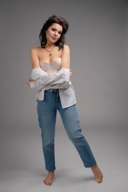 Bir kadın kendine güvenerek poz verir, korse, kot pantolon ve açık renk gömlek giyer. Görünüşü, minimalist bir stüdyoda zarafet ve modern tarzı birleştiriyor..