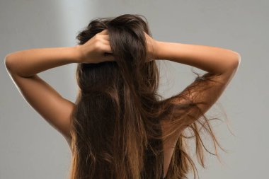 Bir kadın sırtı seyirciye dönük duruyor, iki eliyle uzun saçlarını kaldırıyor. Yumuşak ışık saçlara ve cilde vurgu yapıyor..