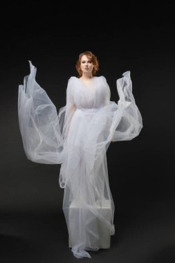 Zarif beyaz iç çamaşırı giymiş çarpıcı bir kadın, akmakta olan kumaşın, somut güzelliğin ve gelin zarafetinin ortasında duruyor..