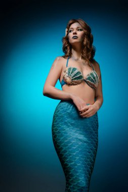 Kendine güvenen bir kadın denizkızı kostümü içinde poz veriyor, vücut sanatı sergiliyor ve mavi arka planda eşsiz bir kuyruğu var..