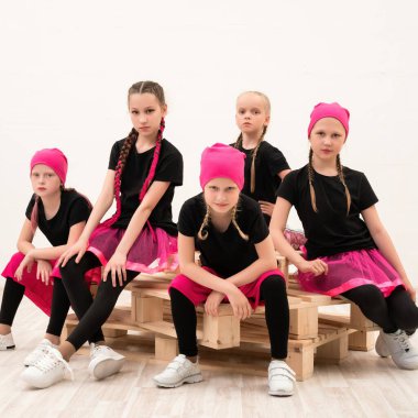 Pembe elbiseli beş kız, ahşap paletlere oturmuş dans kıyafetlerini sergiliyor ve modern stüdyoda çarpıcı pozlar veriyorlar..