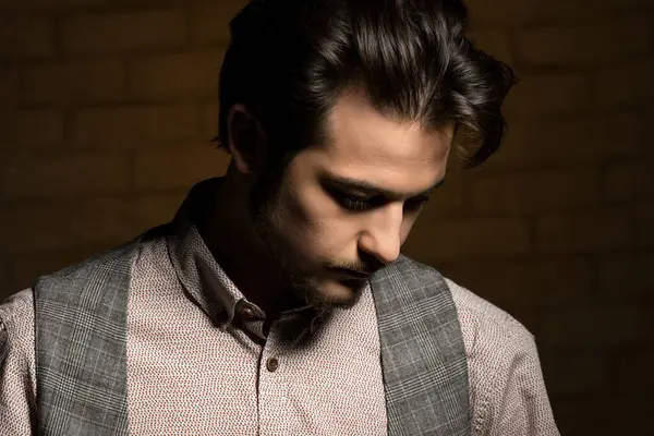 Kalıplı gömlek ve yelek giyen genç bir adam, yumuşak, sıcak bir ışıkla aydınlanan, dokulu bir arka planda yansıtıcı bir ifadeyle aşağı bakar..