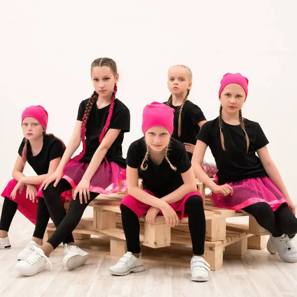 Pembe elbiseli beş kız, ahşap paletlere oturmuş dans kıyafetlerini sergiliyor ve modern stüdyoda çarpıcı pozlar veriyorlar..