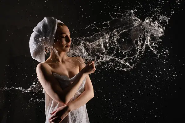 Havluya sarılmış bir kadın karanlık bir alanda neşeyle su sıçratır ve etrafına sanatsal damlalar oluşturur..