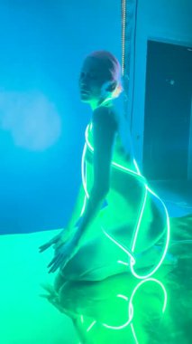 Bir kadın göz kamaştırıcı iç çamaşırlarıyla özgüven ve zarafet yayar, sahne arkasında canlı bir ışık altında poz verir..