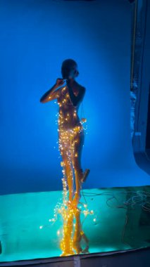 Göz kamaştırıcı bir kadın narin iç çamaşırlarıyla poz verir, güzel bir şekilde parlak ışıklara sarılır, mavi bir arkaplana karşı..