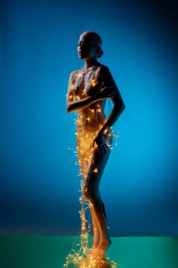 Bir kadın zarif bir şekilde duruyor, yumuşak ışıklarla süslenmiş, sakin bir ortamda güzelliğini parlak mavi bir arka planla ön plana çıkarıyor..