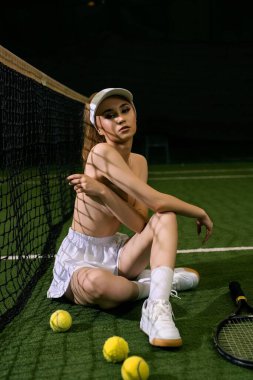 Tenis toplarıyla çevrili bir kadın tenisçi, kortta oturur ve gece antrenmanı sırasında rahat bir poz verir..