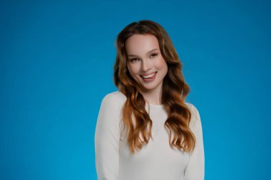 Stüdyoda kendine güvenen bir kadın, parlak bir gülümsemeyle mutluluk saçıyor. Dalgalı saçları, çarpıcı bir mavi arkaplan karşısında poz verirken yüzünü güzelce çerçeveler ve canlandırıcı bir anı yakalar..