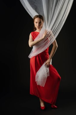 Bir kadın stüdyoda zarif bir şekilde duruyor, çarpıcı kırmızı bir elbise giymiş. Saf kumaşı tutarken, bir dizi duygu sergiliyor. Zarif ve sanatsal bir atmosfer yaratıyor..