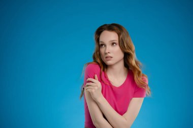 Bir kadın stüdyoda poz veriyor, düşünceli bir bakışla duygularını sergiliyor. Sade pembe bir bluz giyer, parlak mavi arka planla çarpıcı bir kontrast yaratır..