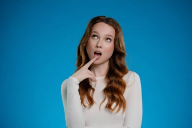 Canlı bir stüdyo arkaplanında, bir kadın neşeli bir ruh hali sergilerken düşünceli bir şekilde parmaklarıyla dudaklarında poz veriyor, izleyicileri etkileyici bakışları ve parlaklığıyla kendine çekiyor..