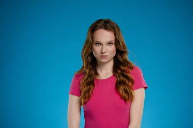 Bir kadın canlı, mavi bir geçmişi olan bir stüdyoda kendine güvenerek duruyor. Dalgalı saçları var ve pembe bir bluz giyiyor. Seyirciyi büyüleyen güçlü ve anlamlı bir duygu sergiliyor..