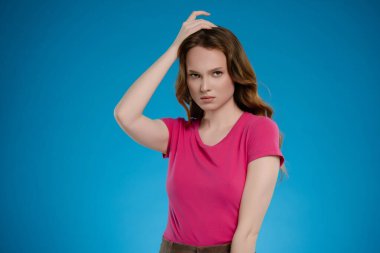 Bir kadın stüdyoda güven içinde duruyor, tek eliyle nazikçe saçına dokunuyor ve güçlü bir duygusal varlık sergiliyor. Parlak mavi arka plan çarpıcı duruşunu güçlendiriyor..