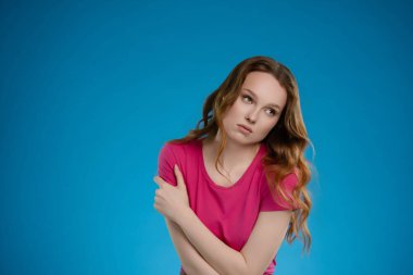 Stüdyoda düşünceli bir şekilde poz veren bir kadın. İfadesi, canlı pembe bluzu ve çarpıcı mavi arkaplanı tarafından vurgulanan duygularının bir karışımını ortaya koyuyor..