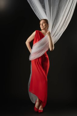 Bir kadın çarpıcı kırmızı elbisesiyle zarif bir şekilde ayakta duruyor, ifadesini derin duygularla ifade ediyor. Stüdyoda örtülmüş hassas kumaşlarla etkileşime geçiyor ve duruşunu sergiliyor..