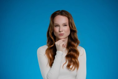 Bir kadın stüdyoda durup düşünceli bir jestle neşeli bir tavır sergiliyor. Uzun, dalgalı saçları parlak mavi arkaplanı tamamlıyor ve göz alıcı bir kontrast oluşturuyor..