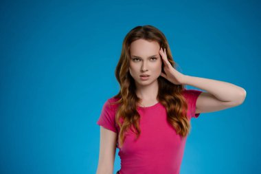 Bir kadın, yoğun duygularını ifade ederken, elini alnına koyduğu parlak bir stüdyoda duruyor. Pembe bir gömlek giyer ve kamera karşısında saçı dökülür..