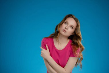 Profesyonel bir stüdyo ortamında, bir kadın düşünceli bir ifadeyle poz verir. Uzun saçları omuzlarının etrafında dalgalanır, parlak mavi arka planla çelişir..