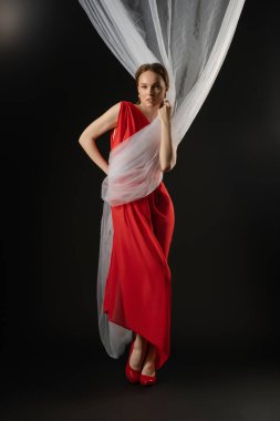 Bir stüdyo ortamında, bir kadın kendinden emin bir şekilde etrafını saran bir kumaş parçasıyla ayakta durur. Kırmızı bir elbise ve topuklu ayakkabı giyerek güçlü bir duygusal varlık gösteriyor..