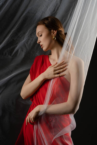 A woman stands gracefully in a studio setting, gently holding a sheer piece of fabric.