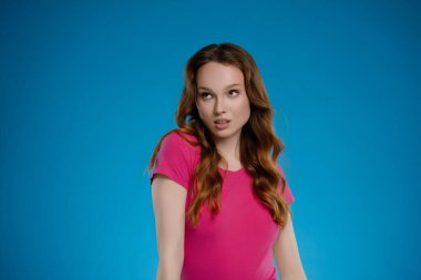 Bir kadın stüdyoda duruyor, açık pembe bir bluz giyerek sanatsal bir anı yakalayan canlı bir mavi arkaplan karşısında neşeli ve duygusal bir ifade sergiliyor..