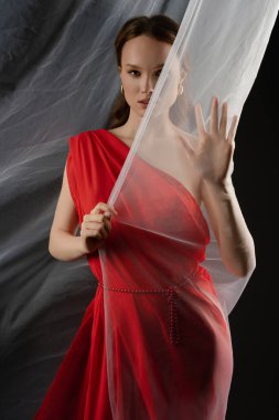 Kontrollü bir stüdyo ortamında canlı kırmızı elbiseli bir kadın incinebilirliğini gösterirken zarifçe poz verir. Eli yarı şeffaf bir kumaştan uzanıyor, güçlü bir duygu sergiliyor..