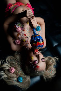 Bir kadın karanlık bir yüzeyde uzanıyor, renkli vücut sanatı ve tatlılarla çevrili, oyuncu sanatlar sergiliyor..