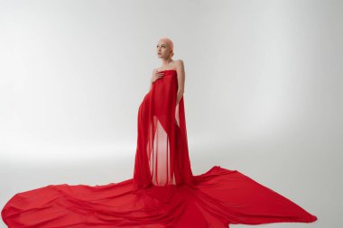 Narin kırmızı kumaşlı bir kadın zarif bir şekilde duruyor, sakin, minimalist bir ortamda kendine güven ve zarafet yayıyor..