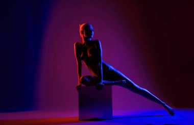 Bir dansçı, canlı kırmızı ve mavi ışıklarla aydınlatılan bir küp üzerinde zarif bir poz verir. Silüetini sanatsal bir şekilde vurgular..