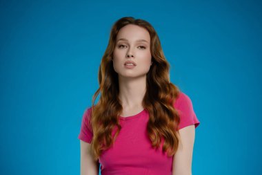 Bir kadın stüdyo ortamında kendine güveni tam bir şekilde duruyor, bir dizi duygu sergiliyor. Uzun, dalgalı saçları var ve mavi arka planla çelişen parlak pembe bir bluzu var..