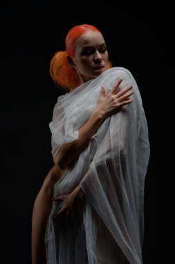 Kızıl saçlı bir kadın saf kumaş içinde zarifçe poz verir. Karanlık bir ortamda ışıldayan güzellik ve kendine güven..