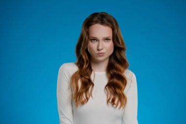 İyi ışıklandırılmış bir stüdyoda, bir kadın canlı mavi bir arkaplana karşı poz veriyor. Yüzündeki ifade, düşünceli bir duyguyu yansıtıyor. Çekimin sanatsal etkisini arttırıyor..