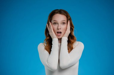 Stüdyoda poz veren bir kadın, elleri yüzünde, şaşırmış bir bakış sergiliyor. Basit mavi arkaplan onun anlamlı özelliklerini ve duygusal görüntüsünü arttırır..