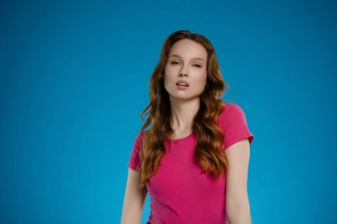 Bir kadın stüdyoda duruyor, özgüven ve duygu yayıyor. Uzun saçları omuzlarının üzerinde dalgalı pembe bir gömlek içinde poz verirken mavi arkaplan karşısında çarpıcı bir kontrastlık oluşturuyor..