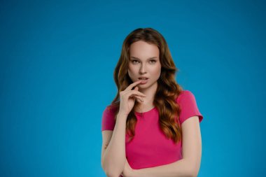 Bir kadın stüdyoda güven içinde duruyor, uzun saçları omuzlarının üzerinden dökülüyor. Kameraya bağlanıyor, düşünceli ve duygusal bir ifade sergiliyor..