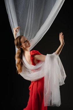 Bir kadın stüdyoda zarif bir şekilde poz veriyor, bir dizi duygu sergiliyor. Çarpıcı kırmızı bir elbise giyiyor ve kompozisyona hareket ve derinlik katan parlak, dalgalı kumaşlarla süsleniyor..