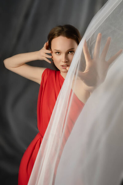 In a studio setting, a woman in a vibrant red dress poses gracefully, using her hands to push aside sheer fabric. Her expression conveys deep emotion, highlighting her artistry and presence.