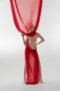 Güzel bir kadın kendine güvenerek duruyor, sadece saf kırmızı kumaşla süslenmiş, yumuşak ışıklı bir çevrede zarafet sergiliyor..