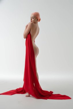 Bir kadın zarif poz verir, kısmen hafif kumaşla örtülmüş, doğal güzelliğini basit bir arka plana vurgular..