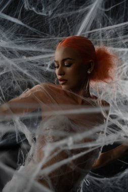 Kızıl saçlı bir kadın, narin bir ağa takılıp, güzelliğini ön plana çıkarıyor ve karmaşık ağın içinde duruyor..